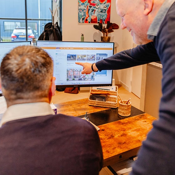 Makelaars staan achter een bureau, één makelaar wijst naar het computerscherm