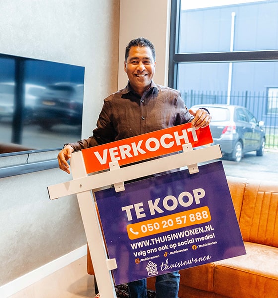 Makelaar staat glimlachend met een verkoopbord in handen